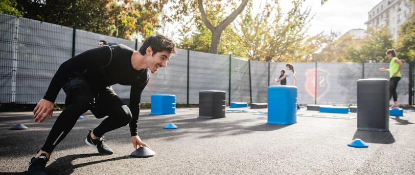 A man in athletic clothing is engaged in an outdoor workout or training session, bent forward while holding a marker or cone. The environment includes other sports equipment such as blue and gray barrels and more cones scattered around. In the background, additional people are seen participating, with trees and a building visible, suggesting a park or recreational area.